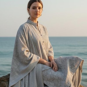 May include: A light grey outfit comprising a long-sleeved tunic with a mandarin collar and matching loose trousers. The ensemble has a subtle floral pattern. The person is sitting on a rock near the sea.