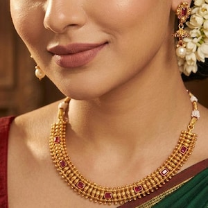 May include: A woman wearing gold jewelry, including a necklace and earrings with red gemstones. The necklace and earrings are gold-toned with a detailed design. The woman has a red bindi on her forehead and is wearing a red top.