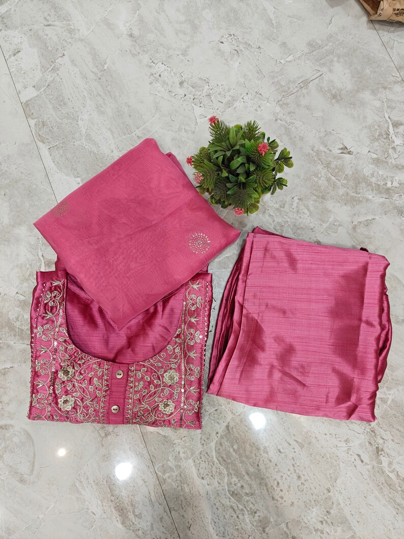May include: A three-piece ensemble in a dusty rose hue. The set includes a tunic with intricate embroidery and sequin detailing, matching pants, and a coordinating scarf. A small potted plant with pink flowers sits nearby.