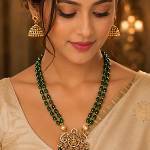 May include: A double-strand necklace featuring smooth, dark green oval beads and a central gold-toned pendant with intricate filigree, small white pearls, and a central deity figure, paired with matching gold-toned bell-shaped jhumka earrings.