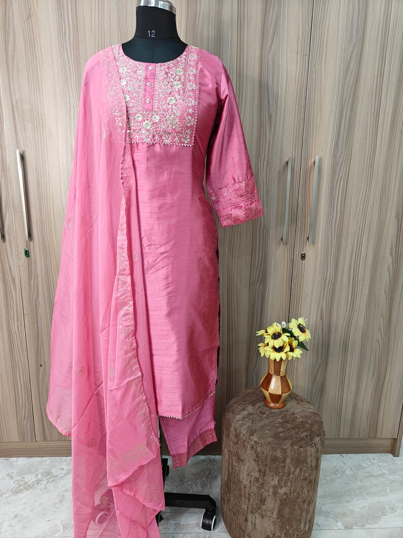 May include: A pink salwar kameez set displayed on a mannequin. The set includes a long-sleeved tunic with embroidered detailing, matching pants, and a sheer dupatta. The fabric has a subtle sheen.