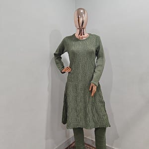 May include: A green knitted sweater dress and matching trousers set. The sweater has a cable knit pattern and a slightly flared skirt. The trousers are fitted and have a straight leg.