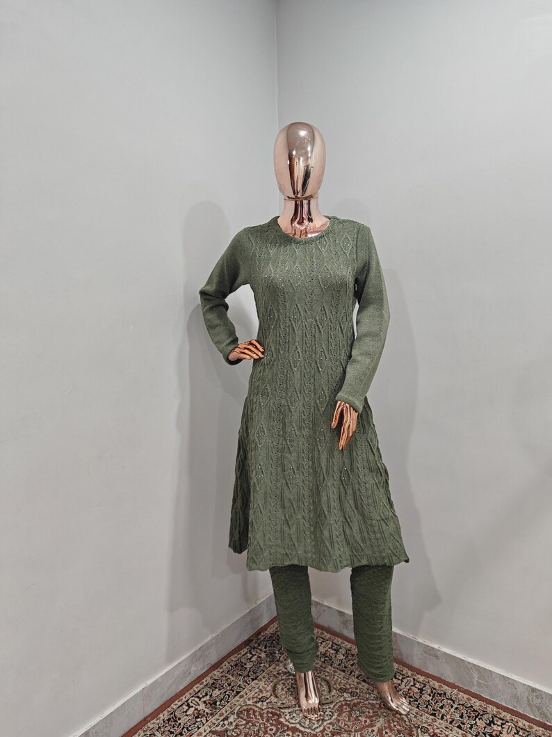 May include: A green knitted sweater dress and matching trousers set. The sweater has a cable knit pattern and a slightly flared skirt. The trousers are fitted and have a straight leg.