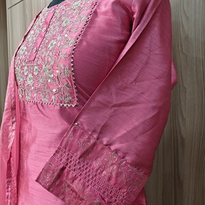 May include: A dusty rose-colored tunic with long sleeves, featuring intricate gold embroidery around the neckline and on the cuffs. The fabric has a subtle sheen, and the design includes a small, decorative trim.