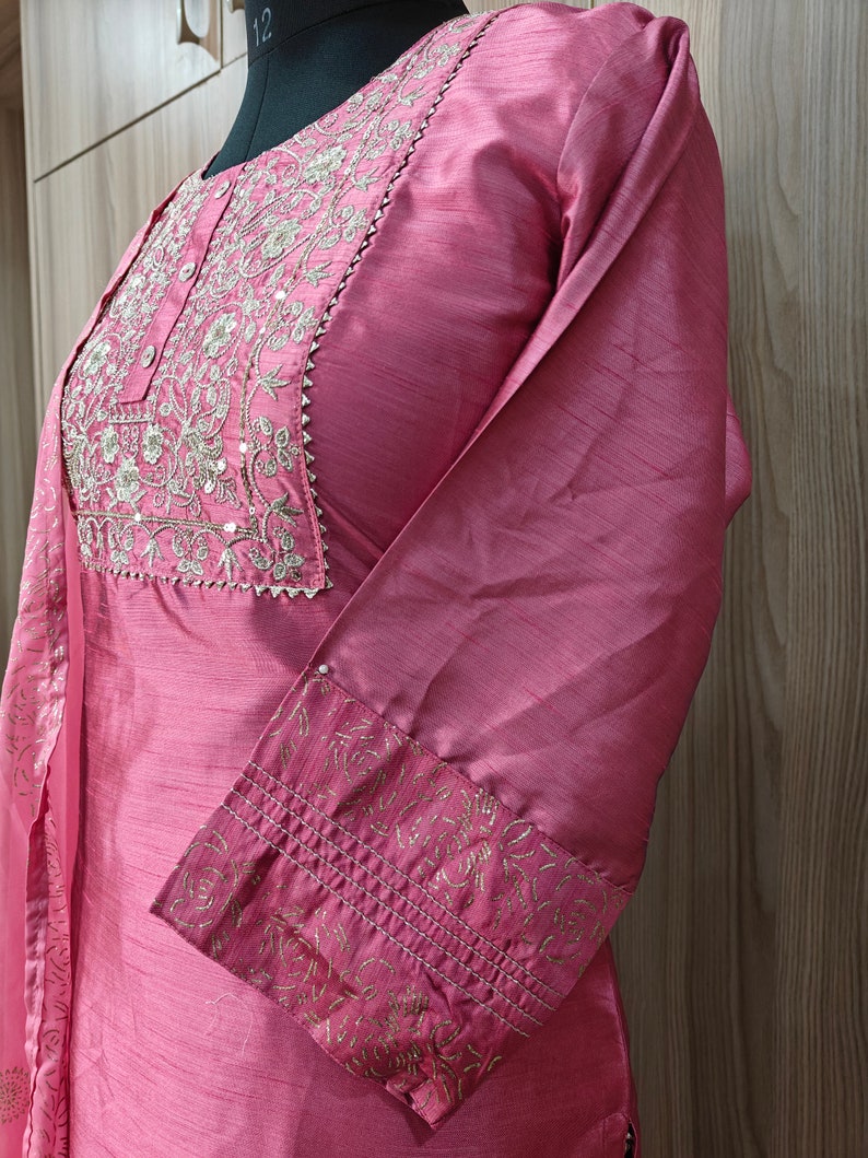 May include: A dusty rose-colored tunic with long sleeves, featuring intricate gold embroidery around the neckline and on the cuffs. The fabric has a subtle sheen, and the design includes a small, decorative trim.