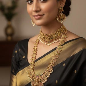 May include: A woman wearing an ornate gold necklace, earrings, and choker set. The jewelry features intricate designs with red, green, and white accents. She is wearing a black sari with a gold border.