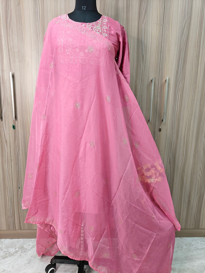 May include: A light pink, long-sleeved dress with intricate embroidery around the neckline and scattered across the fabric. The dress features a flowing, layered design, and is displayed on a mannequin. The fabric appears lightweight and semi-sheer.