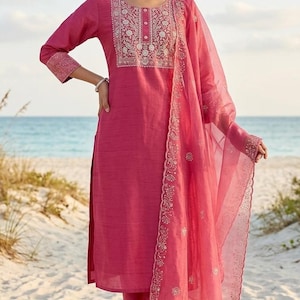 May include: A magenta-colored Indian salwar kameez ensemble. The outfit includes a long-sleeved tunic with embroidered detailing, matching pants, and a sheer dupatta. The outfit is worn on a sandy beach with the ocean in the background.