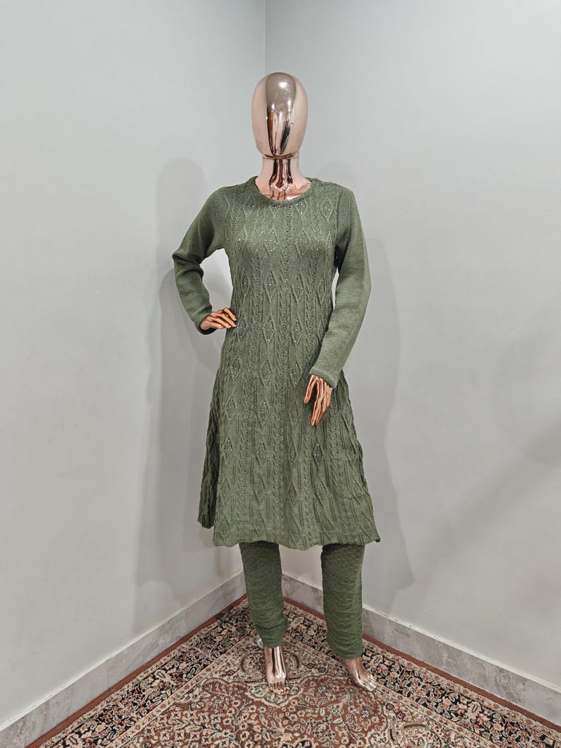 May include: A long-sleeved, green knitted sweater dress with a diamond pattern and a matching pair of trousers. The outfit is displayed on a mannequin.