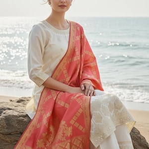 May include: A woman wearing a cream-colored embroidered kurta and white pants, with a coral and gold patterned dupatta. The outfit is traditional Indian wear, suitable for festive occasions. The background shows the ocean.