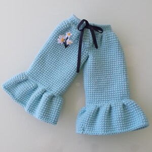 May include: Light blue crochet pants with a ruffled hem and a dark blue ribbon tie. The pants feature a small embroidered floral design near the waist. The crochet stitch creates a textured appearance.
