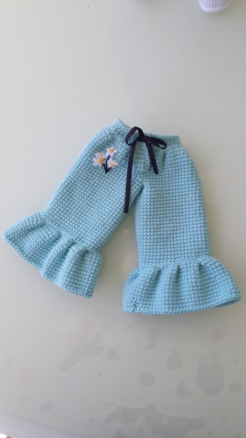 May include: Light blue crochet pants with a ruffled hem and a dark blue ribbon tie. The pants feature a small embroidered floral design near the waist. The crochet stitch creates a textured appearance.