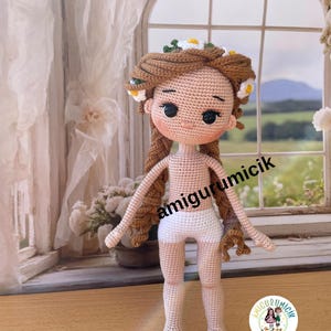 May include: A handmade crochet doll with brown hair adorned with white and yellow flowers. The doll has a light skin tone, black eyes, and is wearing white shorts. The text "amigurumicik" is visible on the image.