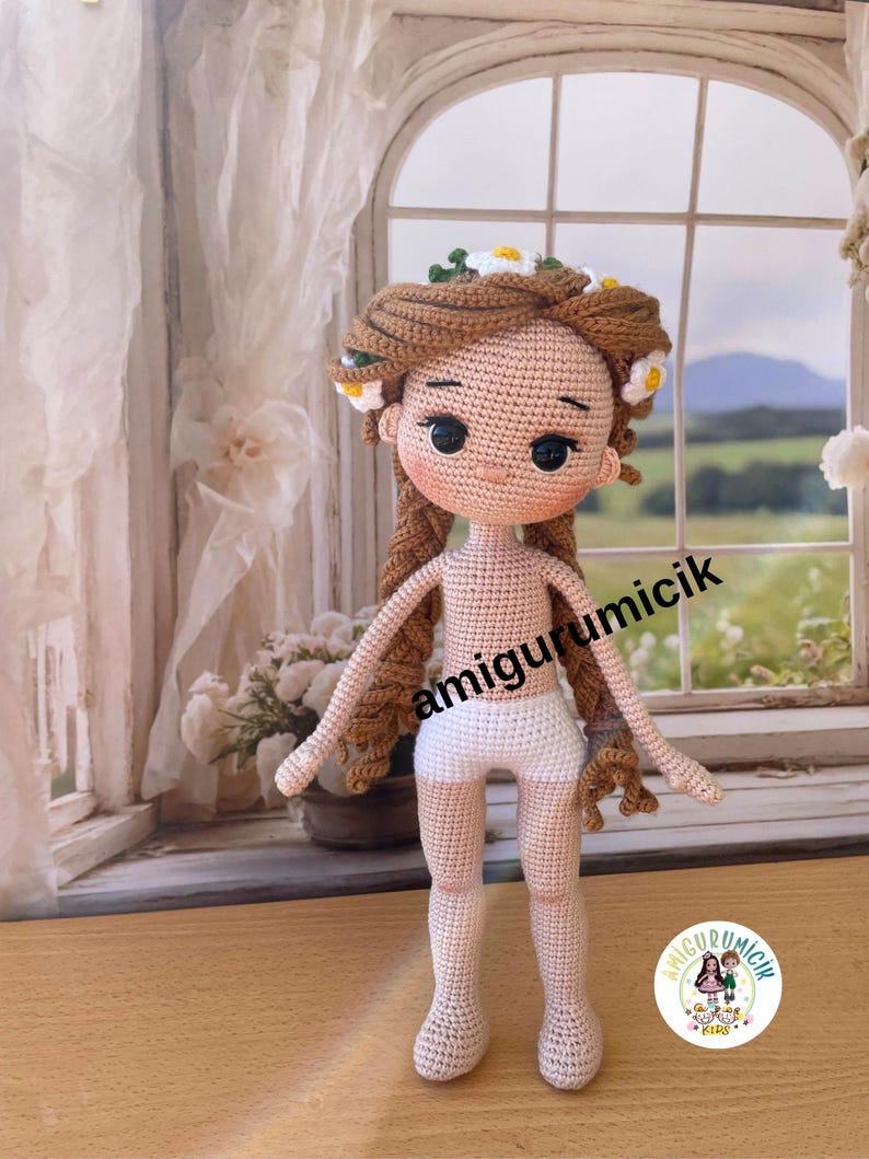 May include: A handmade crochet doll with brown hair adorned with white and yellow flowers. The doll has a light skin tone, black eyes, and is wearing white shorts. The text "amigurumicik" is visible on the image.
