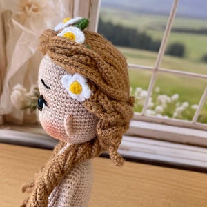 May include: A handmade crochet doll with brown hair styled in braids and adorned with white and yellow daisy accents. The doll has a light skin tone, rosy cheeks, and is wearing a light-colored dress. The doll is positioned in front of a window with a scenic view.