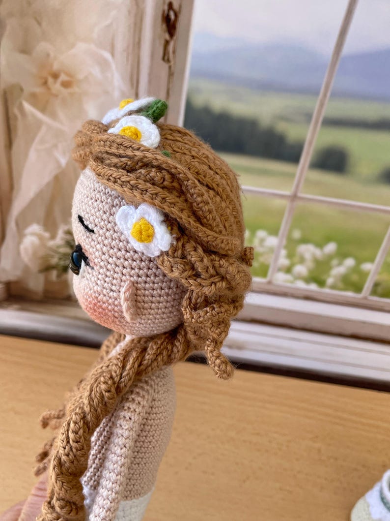 May include: A handmade crochet doll with brown hair styled in braids and adorned with white and yellow daisy accents. The doll has a light skin tone, rosy cheeks, and is wearing a light-colored dress. The doll is positioned in front of a window with a scenic view.