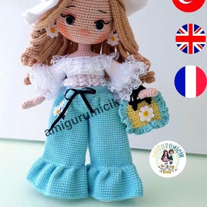 Peut inclure: Poupée crochetée faite à la main, vêtue d'un chapeau blanc, d'un haut et d'un pantalon bleu. La poupée a des cheveux blonds, des boucles d'oreilles marguerites et un petit sac à main jaune. Le texte "amigurumicik" est visible. Objet artisanal.