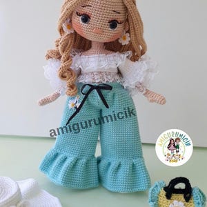 May include: A handmade crochet doll with blonde curly hair, wearing a white top, light blue pants, and daisy earrings. Accessories include a matching hat and purse. The doll's pants have the text "amigurumicik".