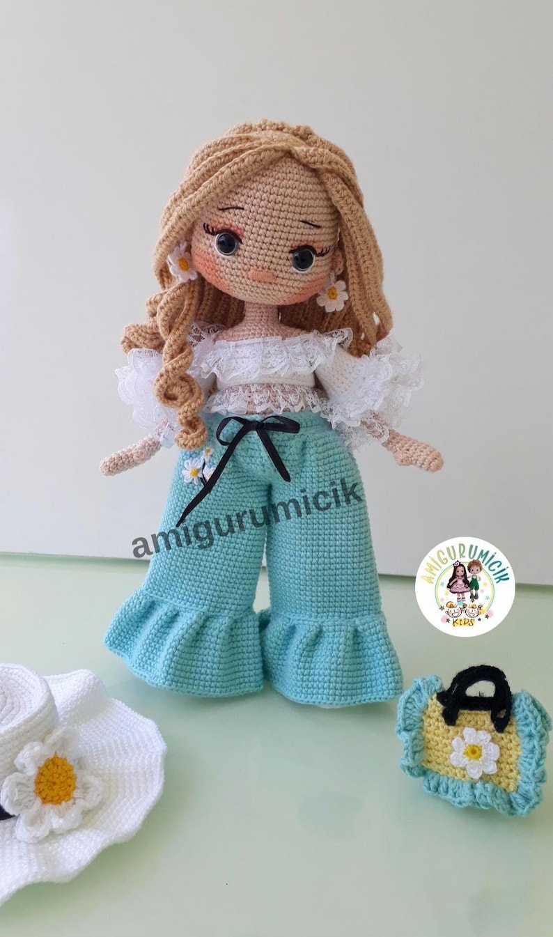 May include: A handmade crochet doll with blonde curly hair, wearing a white top, light blue pants, and daisy earrings. Accessories include a matching hat and purse. The doll's pants have the text "amigurumicik".