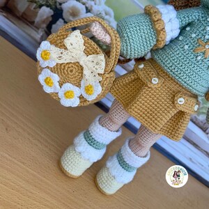 May include: A handmade crochet doll with a yellow skirt, mint green jacket, and white boots. The doll holds a small basket decorated with white daisies and a cream-colored bow. The doll's outfit features button details.