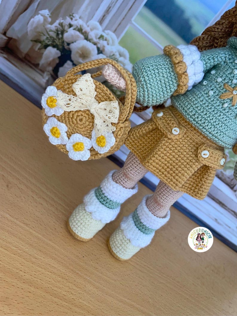 May include: A handmade crochet doll with a yellow skirt, mint green jacket, and white boots. The doll holds a small basket decorated with white daisies and a cream-colored bow. The doll's outfit features button details.