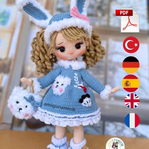 Könnte beinhalten: Handgemachte Häkelpuppe in einem blauen Kleid mit weißer Bordüre und passendem Hut mit Hasenohren. Die Puppe hat lockiges Haar und hält eine weiße Handtasche. Das Outfit der Puppe hat weiße Akzente und eine rosa Schleife.