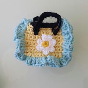 May include: Handmade crochet bib in yellow with a light blue border. The bib features a black strap and a white daisy with a yellow center. The bib is designed for babies and toddlers.