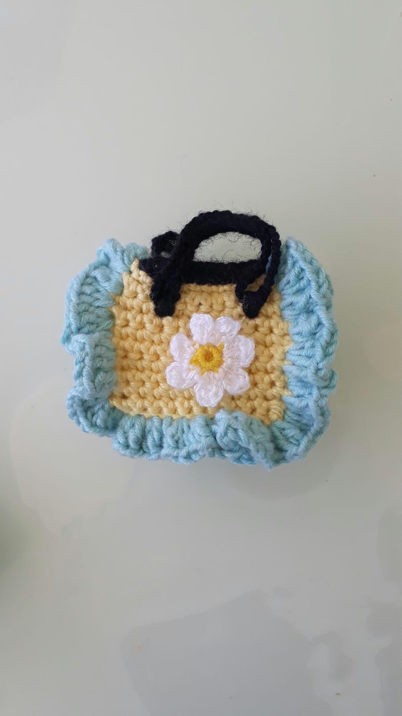 May include: Handmade crochet bib in yellow with a light blue border. The bib features a black strap and a white daisy with a yellow center. The bib is designed for babies and toddlers.
