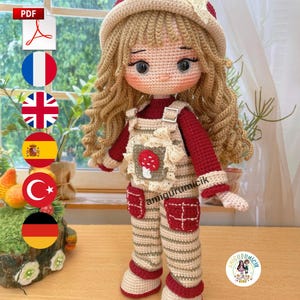 Peut inclure: Poupée au crochet faite à la main avec de longs cheveux blonds bouclés, vêtue d'une chemise rouge à manches longues et d'une salopette rayée avec des accents rouges. La poupée a un chapeau beige avec un nœud et une application de champignon. La poupée mesure environ 30 cm de haut.
