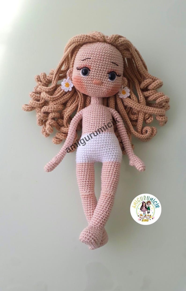 May include: A handmade crochet doll with light brown curly hair and a fair skin tone. The doll has a white diaper and small daisy earrings. The doll's arms and legs are extended. The doll is a handmade amigurumi.