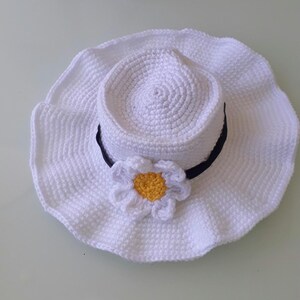 May include: A white crochet sun hat with a wide brim and a black band. A white daisy with a yellow center is attached to the hat. The hat is handmade and appears to be for a doll or small child.