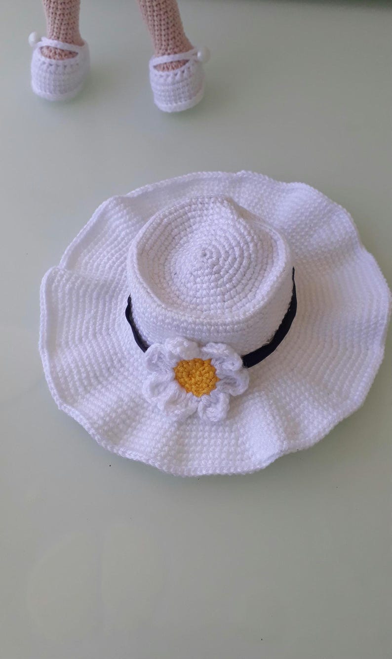May include: A white crochet sun hat with a wide brim and a black band. A white daisy with a yellow center is attached to the hat. The hat is handmade and appears to be for a doll or small child.