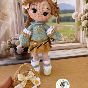 May include: A handmade crochet doll with long brown hair adorned with white and yellow daisy accents. The doll wears a green sweater, a mustard-colored skirt, and white boots. A small basket with daisy embellishments and a bow is also present.