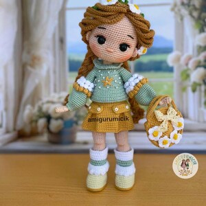 May include: A handmade crochet doll with brown hair, wearing a green sweater, yellow skirt, and white boots. The doll has a daisy crown and carries a small purse decorated with daisies. The doll is standing in front of a window.