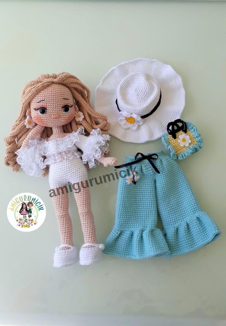 May include: A handmade crochet doll with long blonde hair, wearing a white dress with lace trim and white shoes. Accessories include a white hat, blue pants, and a small purse, all adorned with daisy accents. The doll is approximately 10 inches tall.