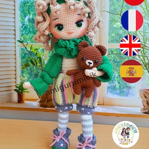 以下が含まれることがあります： 手作りのかぎ針編み人形。カールした髪、緑の目、緑のスカーフがあり、小さな茶色のテディベアを持っています。人形はストライプの服とピンクのリボンが付いた青い靴を履いています。「amigurumicik」の文字が見えます。