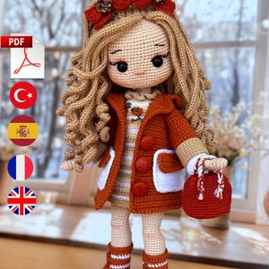 Puede incluir: Muñeca de ganchillo hecha a mano con pelo largo y rubio ondulado y un lazo rojo. La muñeca lleva un abrigo color óxido con ribete blanco, un vestido a rayas y botas a juego. Sostiene un pequeño bolso rojo.