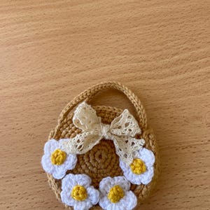 May include: A crocheted, tan-colored purse with a handle, adorned with white daisy-like flowers with yellow centers. A cream-colored, lacy bow is attached to the top of the purse. The purse is round and has a woven texture.