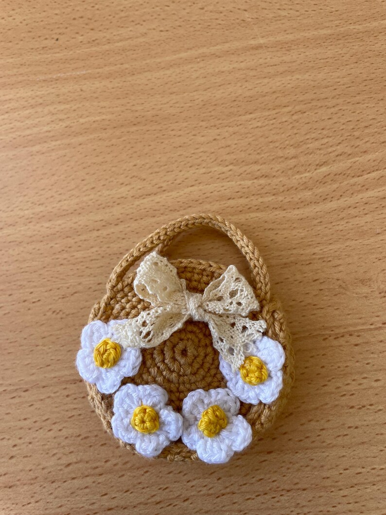 May include: A crocheted, tan-colored purse with a handle, adorned with white daisy-like flowers with yellow centers. A cream-colored, lacy bow is attached to the top of the purse. The purse is round and has a woven texture.