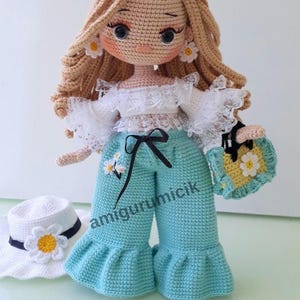 May include: A handmade crochet doll with blonde hair, wearing a white top, turquoise pants, and a matching purse. The doll has daisy earrings and a hat with a daisy. The doll's pants have a black bow. The text "amigurumicik" is on the pants.
