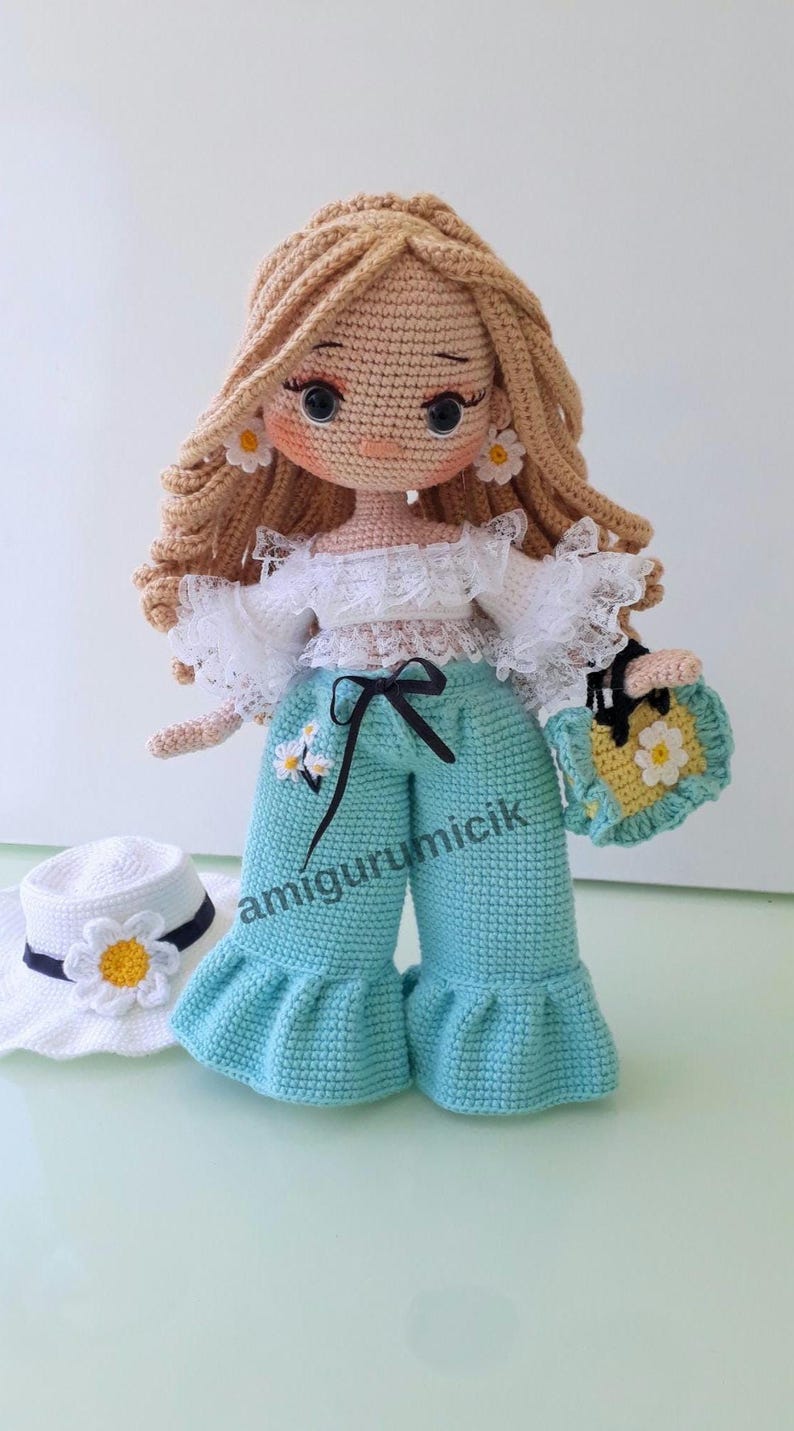 May include: A handmade crochet doll with blonde hair, wearing a white top, turquoise pants, and a matching purse. The doll has daisy earrings and a hat with a daisy. The doll's pants have a black bow. The text "amigurumicik" is on the pants.