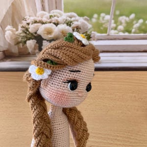 May include: A handmade crochet doll with brown braided hair, adorned with a white and yellow daisy. The doll has a light skin tone, black eyes, and is wearing a white diaper. The doll's hair is styled with a flower crown.