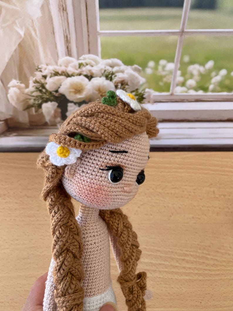 May include: A handmade crochet doll with brown braided hair, adorned with a white and yellow daisy. The doll has a light skin tone, black eyes, and is wearing a white diaper. The doll's hair is styled with a flower crown.