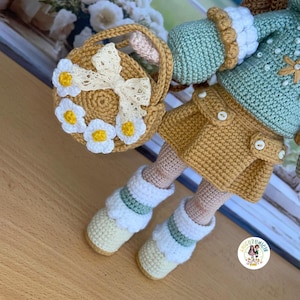 May include: A handmade crochet doll with a yellow skirt, mint green jacket, and white boots. The doll holds a small basket decorated with white daisies and a cream-colored bow. The doll's outfit features button details.