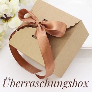 Könnte beinhalten: Eine braune Geschenkbox aus Pappe mit einem gewellten Rand und einem braunen Satinband, das zu einer Schleife gebunden ist. Die Box ist mit "Überraschungsbox" beschriftet, was im Deutschen "Überraschungsbox" bedeutet.