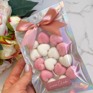 May include: A clear iridescent plastic bag with a pink ribbon tied in a bow. The bag contains pink and white heart-shaped wax melts. The bag has a label that says "Guava Celada".