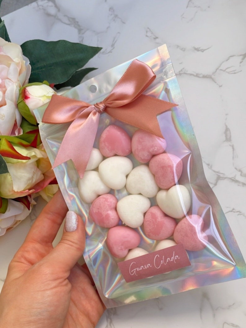 May include: A clear iridescent plastic bag with a pink ribbon tied in a bow. The bag contains pink and white heart-shaped wax melts. The bag has a label that says "Guava Celada".