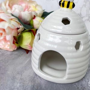 Könnte beinhalten: Weißer Keramikkernwärmer in Bienenstockform mit einer Biene oben drauf. Der Wärmer hat eine runde Öffnung vorne und ein kleines Loch oben.