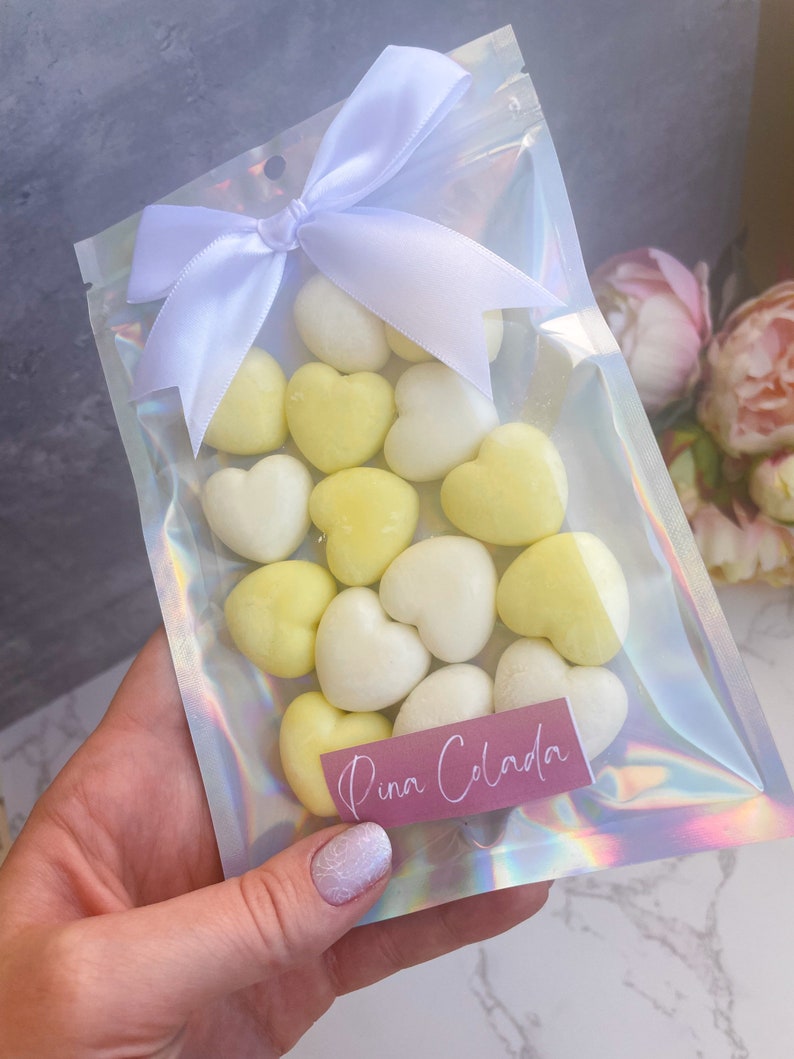 May include: A clear plastic bag with a white bow tied at the top. The bag contains a variety of heart-shaped wax melts in white and yellow. The bag has a pink label that says "Pina Colada".