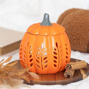 Könnte beinhalten: Ein orangefarbener Keramik-Wärmer in Kürbisform mit grauem Stiel und weizenförmigen Aussparungen. Der Wärmer steht auf einem Holztablett mit Zimtstangen, ideal für die Herbstdekoration. Der Kürbiswärmer ist etwa 15 cm hoch.